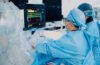 Surgeons in blue scrubs and masks perform an operation, monitoring vital signs on screens nearby.