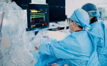 Surgeons in blue scrubs and masks perform an operation, monitoring vital signs on screens nearby.