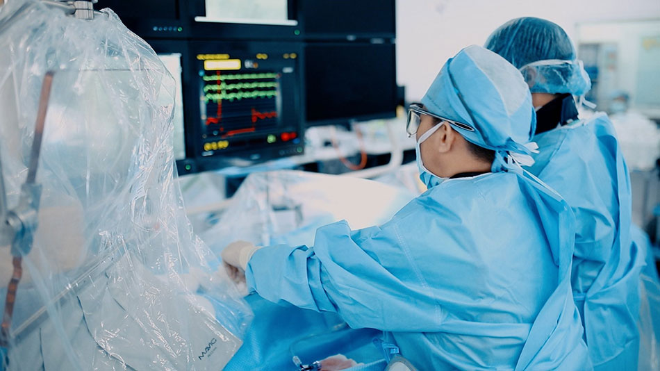 Surgeons in blue scrubs and masks perform an operation, monitoring vital signs on screens nearby.