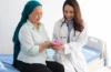 Doctor in a white coat hands a pink object to a patient wearing a teal hijab at a hospital bed.