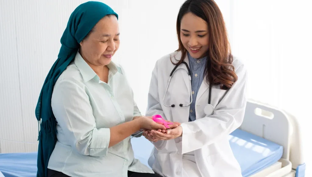 Doctor in a white coat hands a pink object to a patient wearing a teal hijab at a hospital bed.