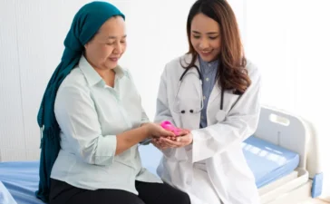 Doctor in a white coat hands a pink object to a patient wearing a teal hijab at a hospital bed.