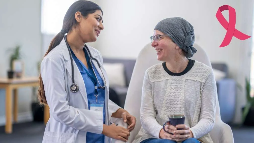 Female doctor in a white coat chats with a cancer patient wearing a headscarf; pink breast cancer ribbon in the corner.